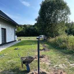 Usługi brukarskie i Ogrodzenia . - Etap montażu ogrodzenia panelowego: wkopane słupki wzdłuż wytyczonej linii, taczka z zaprawą, pracownik w tle. Widok na posesję z domem i zielenią.