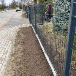 Usługi brukarskie i Ogrodzenia . - Szary panelowy płot z betonową podmurówką wzdłuż chodnika. W tle osoba z taczką i zielone drzewa. Ziemia przygotowana pod nasadzenia. Grabie leżą na ziemi.