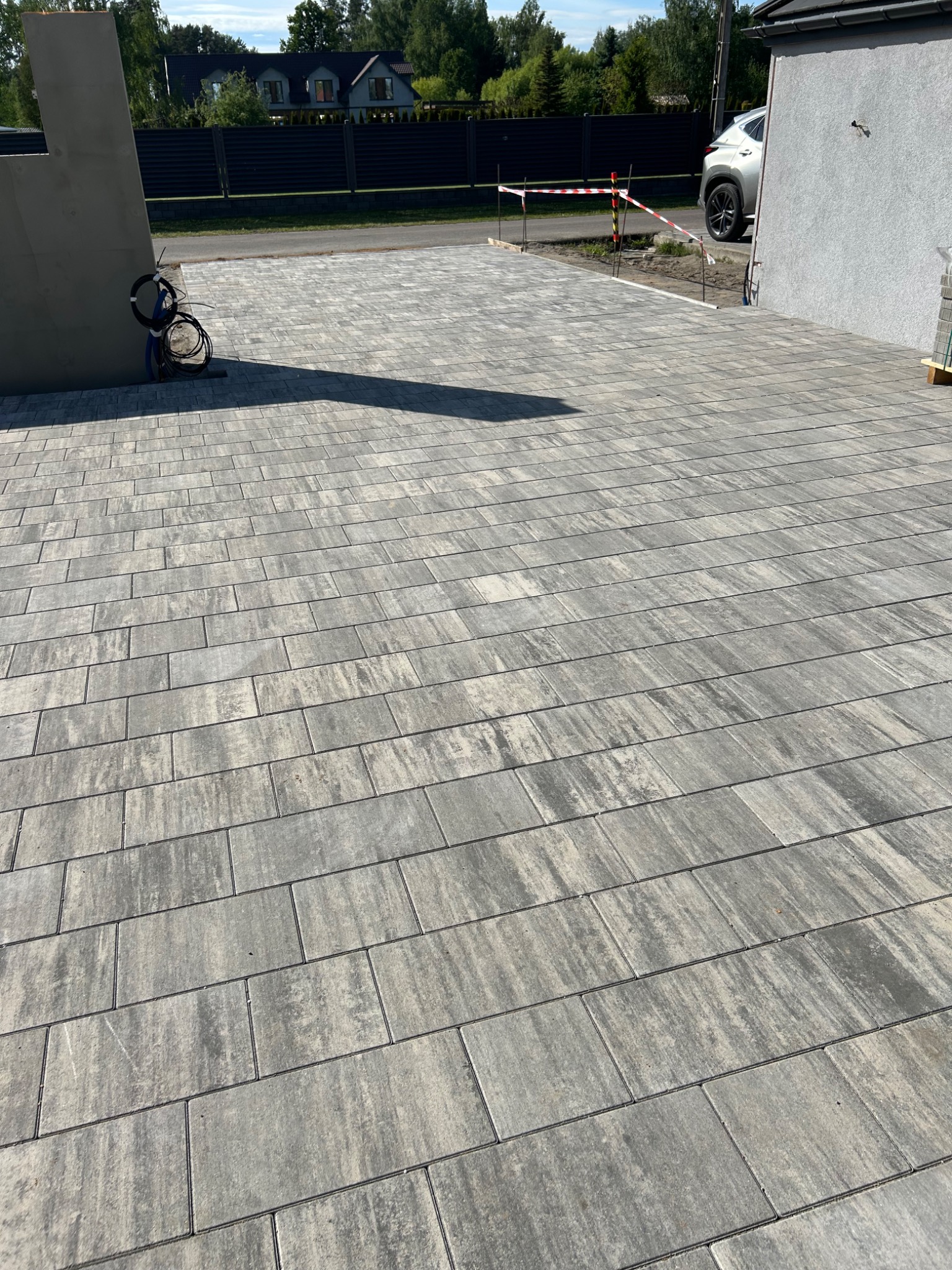 Ułożona kostka brukowa w odcieniach szarości, tworząca wzór prostokątów. W tle widoczny fragment budynku i ogrodzenie, całość w słoneczny dzień.