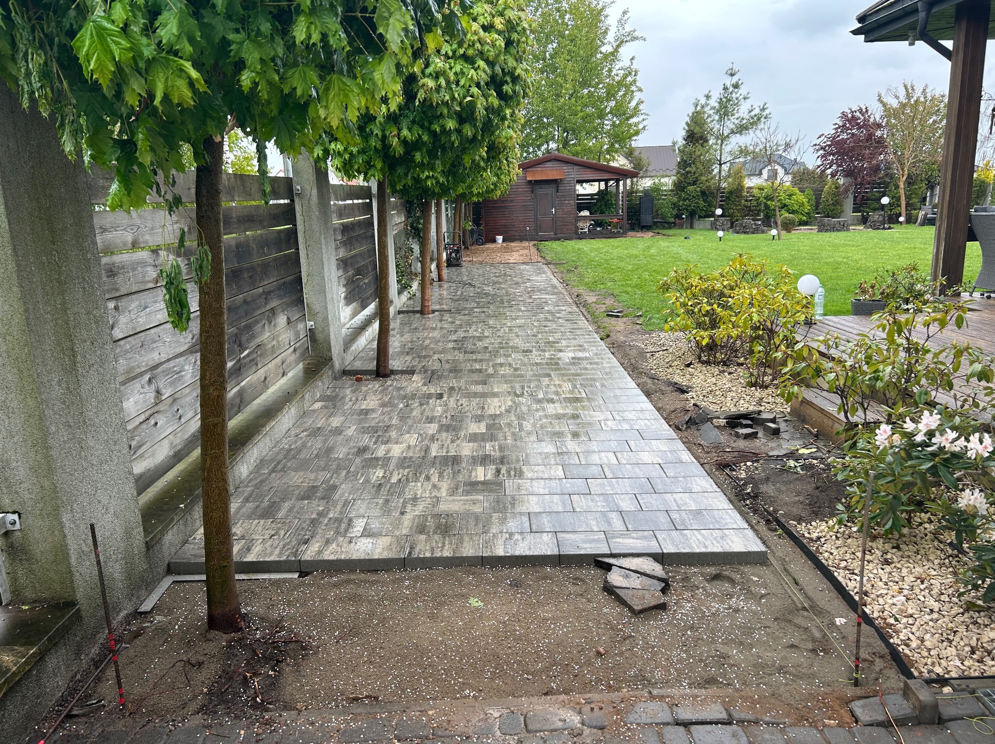 Układanie szarej kostki brukowej na ścieżce w ogrodzie, widoczne drzewa i elementy krajobrazu. Częściowo ukończona praca, z ziemią przygotowaną pod dalsze etapy.