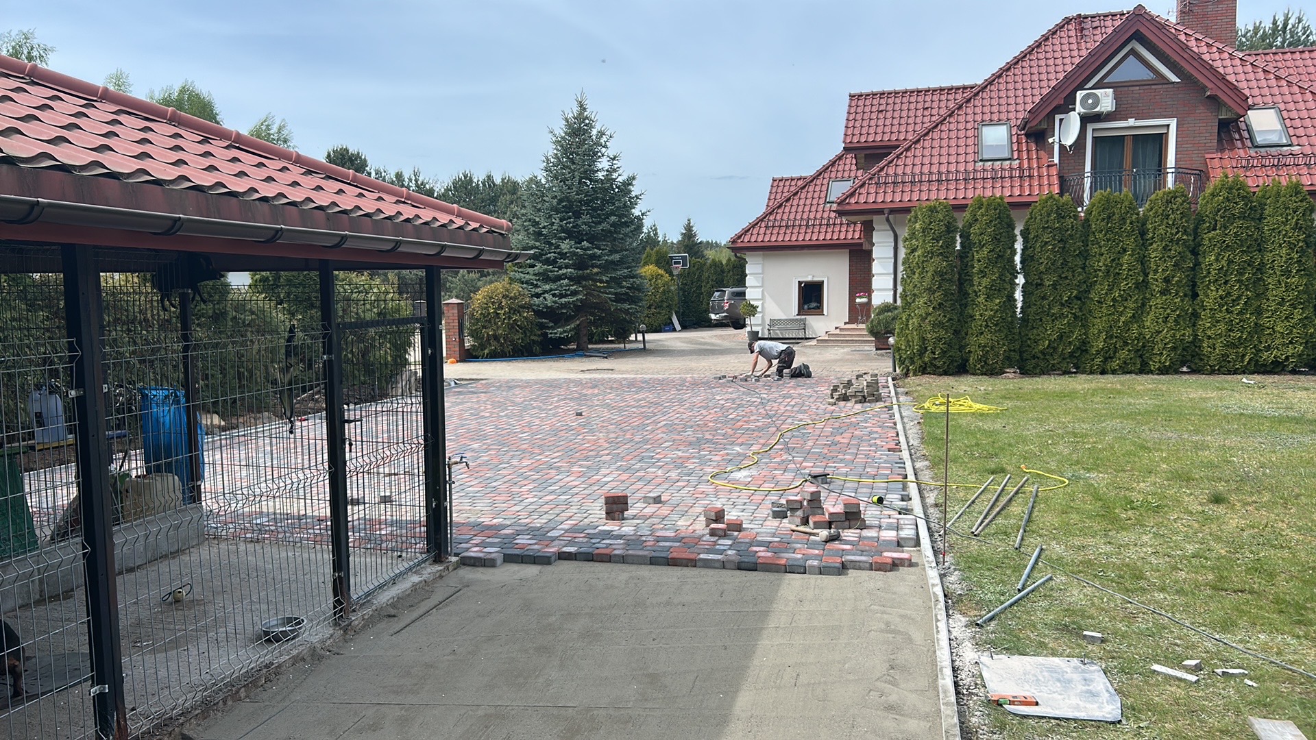Układanie wielobarwnej kostki brukowej na podjeździe domu jednorodzinnego. Widoczny fragment ułożonej nawierzchni i pracujący mężczyzna. W tle dom z czerwoną dachówką.