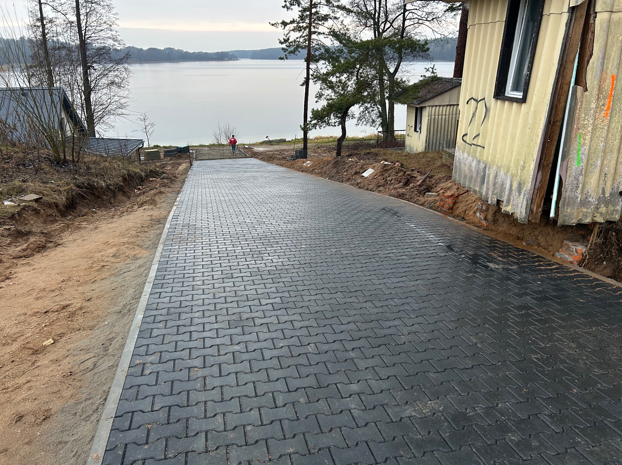 Ułożona, czarna kostka brukowa prowadząca w kierunku jeziora. Po bokach widoczne ziemne skarpy i budynki. W oddali osoba w czerwonej kurtce.