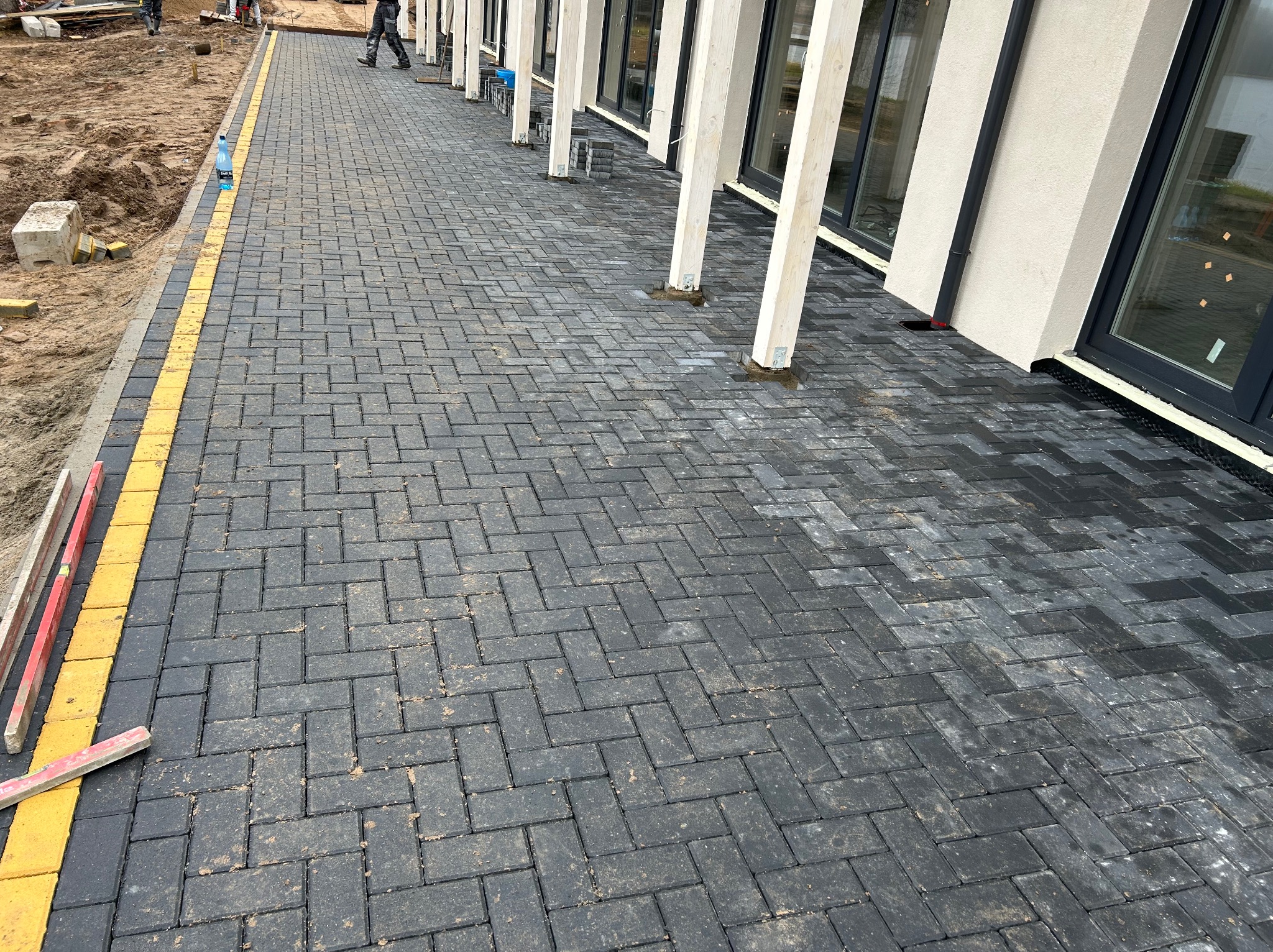 Ułożona, grafitowa kostka brukowa w jodełkę, obrzeże z żółtej kostki. Częściowo widoczne narzędzia i budynek z białymi kolumnami. Świeżo po deszczu.