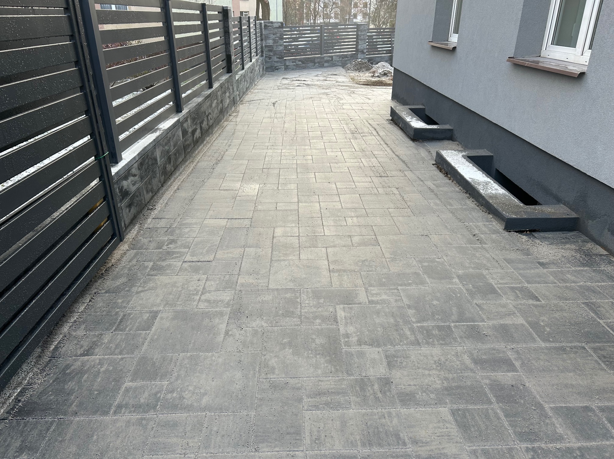 Ułożona kostka brukowa w Augustowie, tworząca ścieżkę wzdłuż nowoczesnego ogrodzenia panelowego z podmurówką. Widoczne okna budynku w tle.