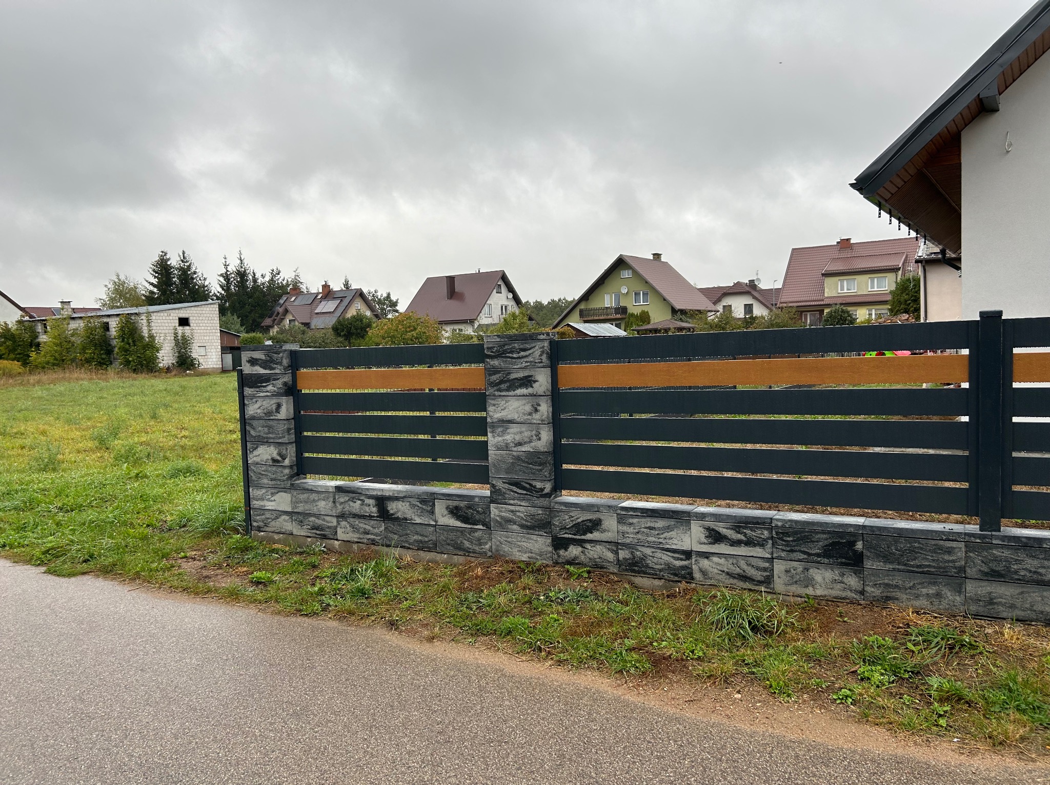 Nowoczesne ogrodzenie z elementami betonowymi i drewnianymi, w tle zabudowa jednorodzinna, pochmurne niebo. Widok z poziomu ulicy.