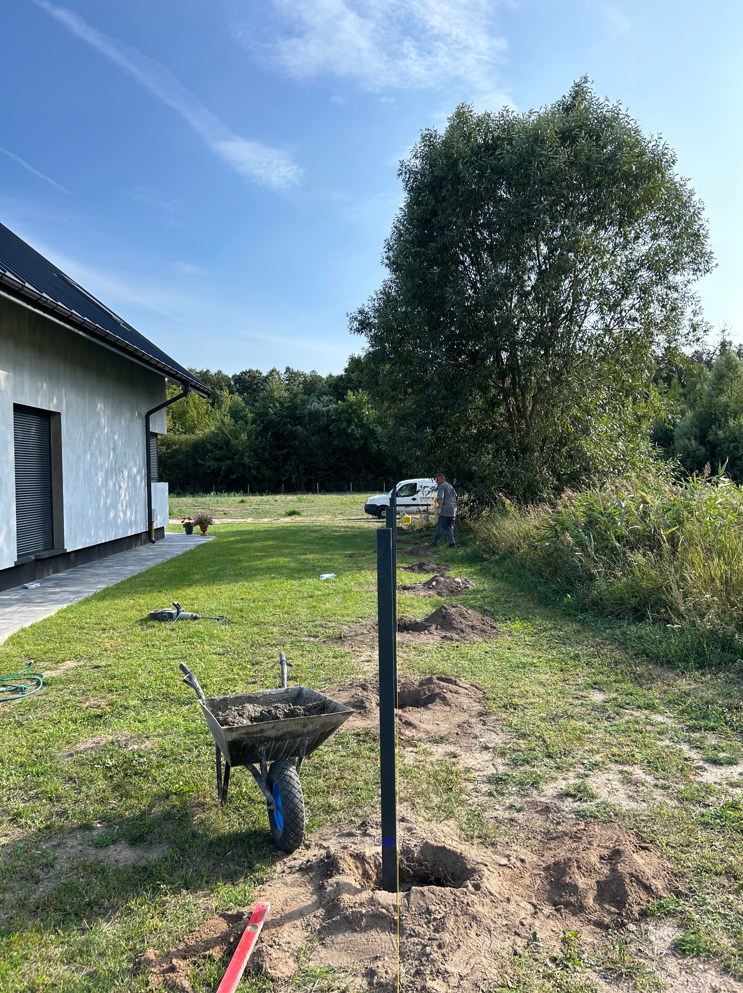 Etap montażu ogrodzenia panelowego: wkopane słupki wzdłuż wytyczonej linii, taczka z zaprawą, pracownik w tle. Widok na posesję z domem i zielenią.