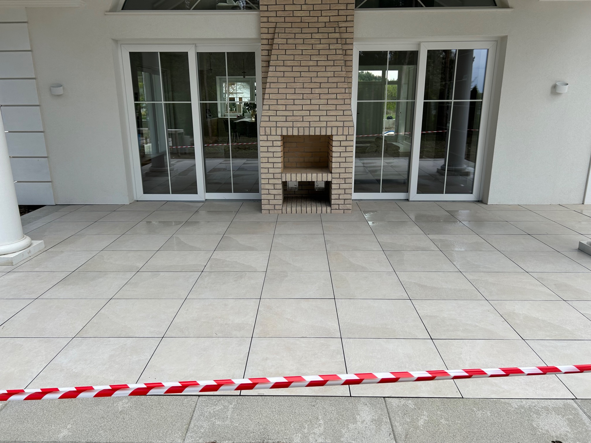 Jasne płytki tarasowe z ceglanym kominkiem zewnętrznym, oddzielone taśmą ostrzegawczą. Widoczne przesuwne drzwi tarasowe i fragment kolumny.