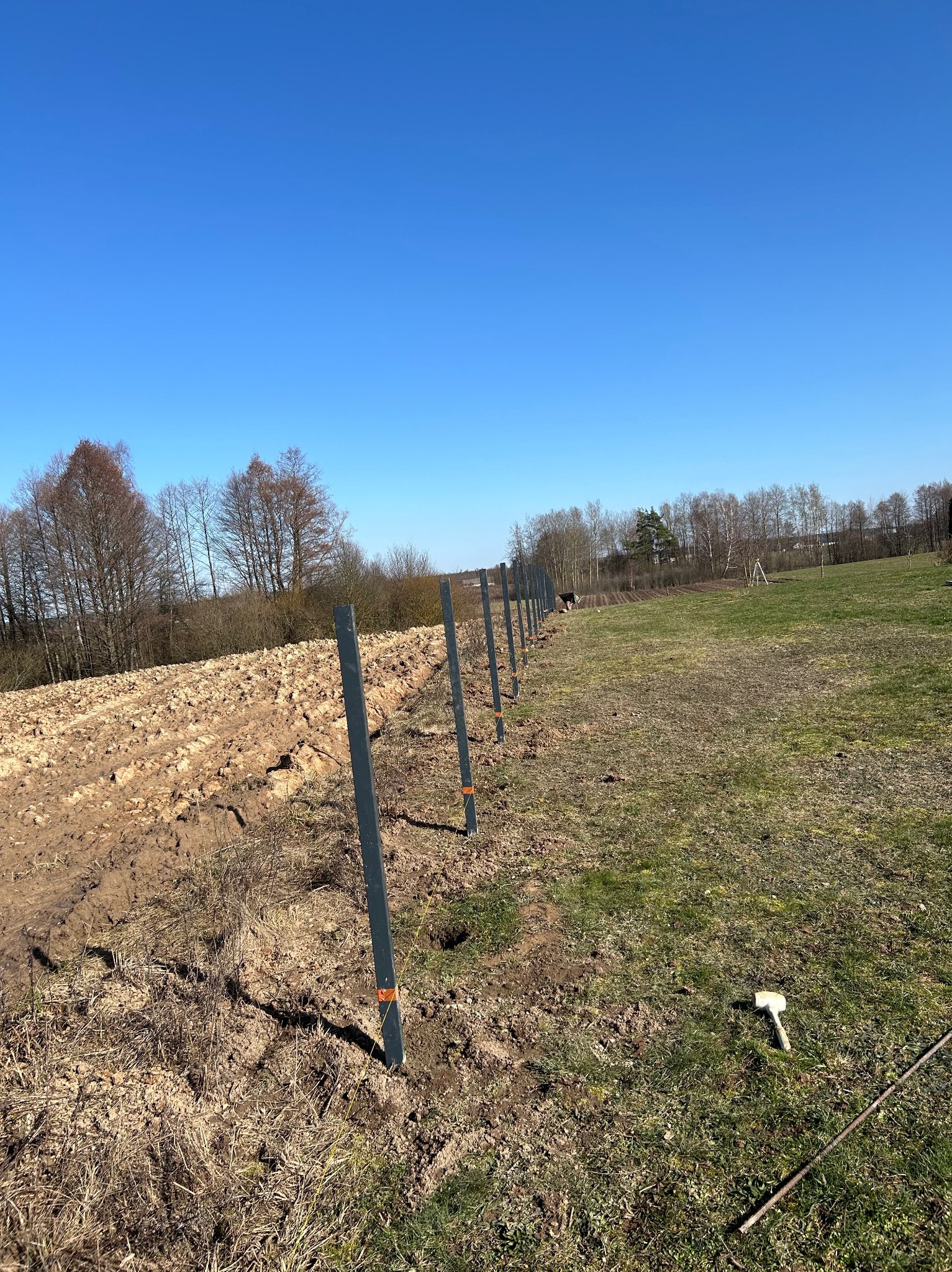 Rząd metalowych słupków ogrodzeniowych, częściowo zamontowanych w ziemi, oddziela zaorane pole od trawnika pod błękitnym niebem. W tle widoczne drzewa i narzędzie pracy.
