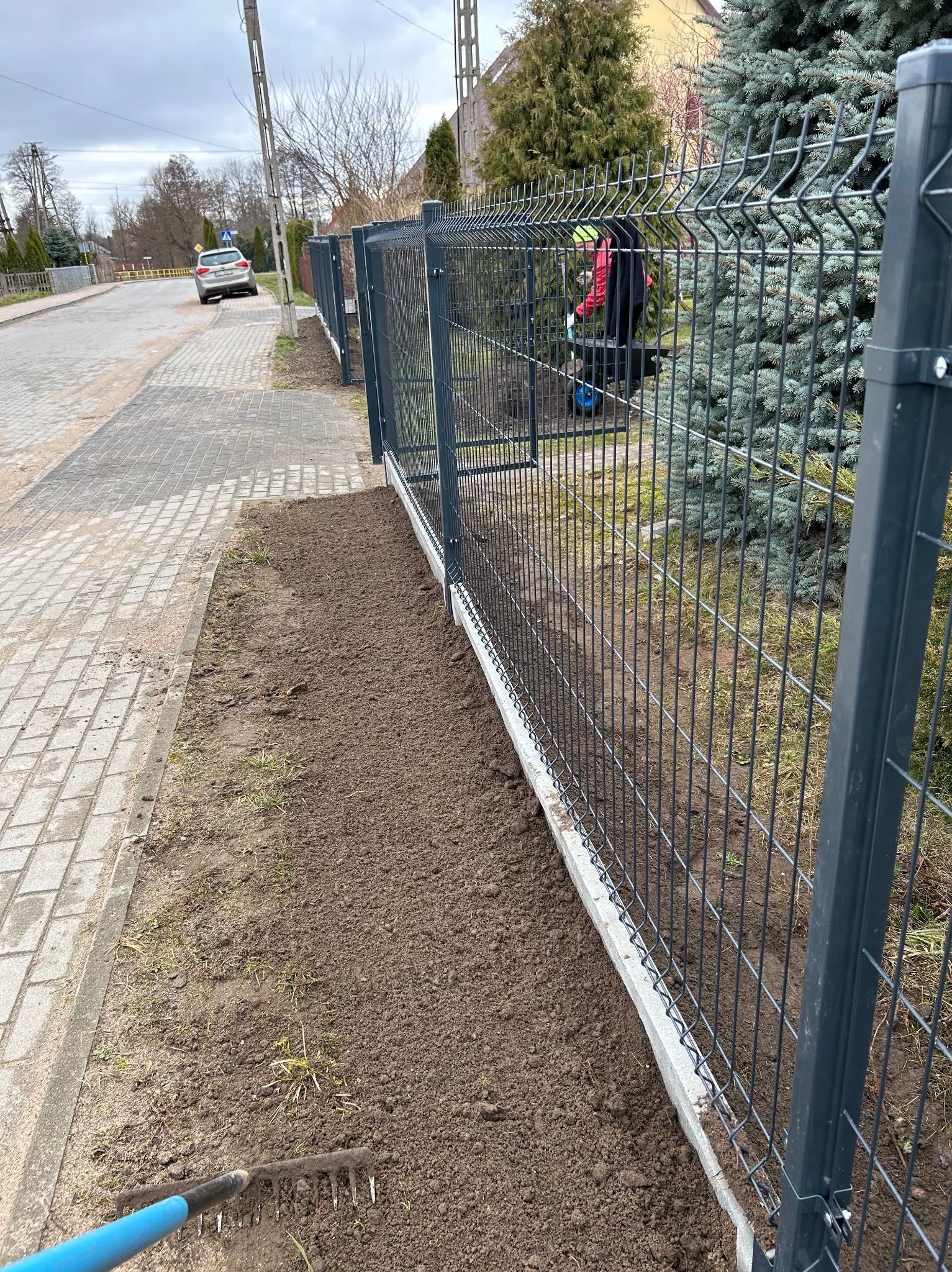 Szary panelowy płot z betonową podmurówką wzdłuż chodnika. W tle osoba z taczką i zielone drzewa. Ziemia przygotowana pod nasadzenia. Grabie leżą na ziemi.