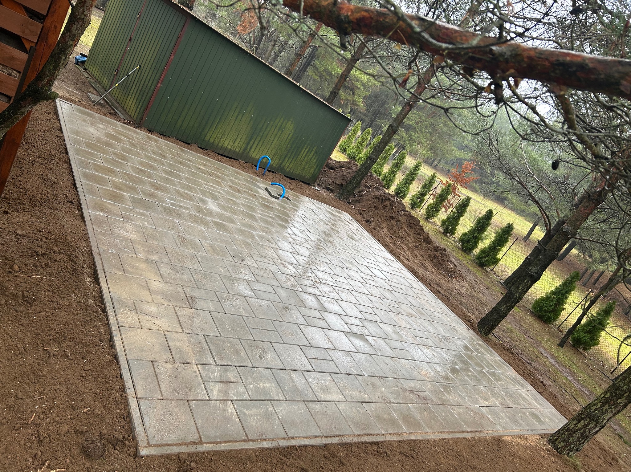 Ułożona, mokra kostka brukowa w kształcie prostokąta na ziemi, obok zielony blaszak i drzewa iglaste. Widok z góry, lekko pod kątem.