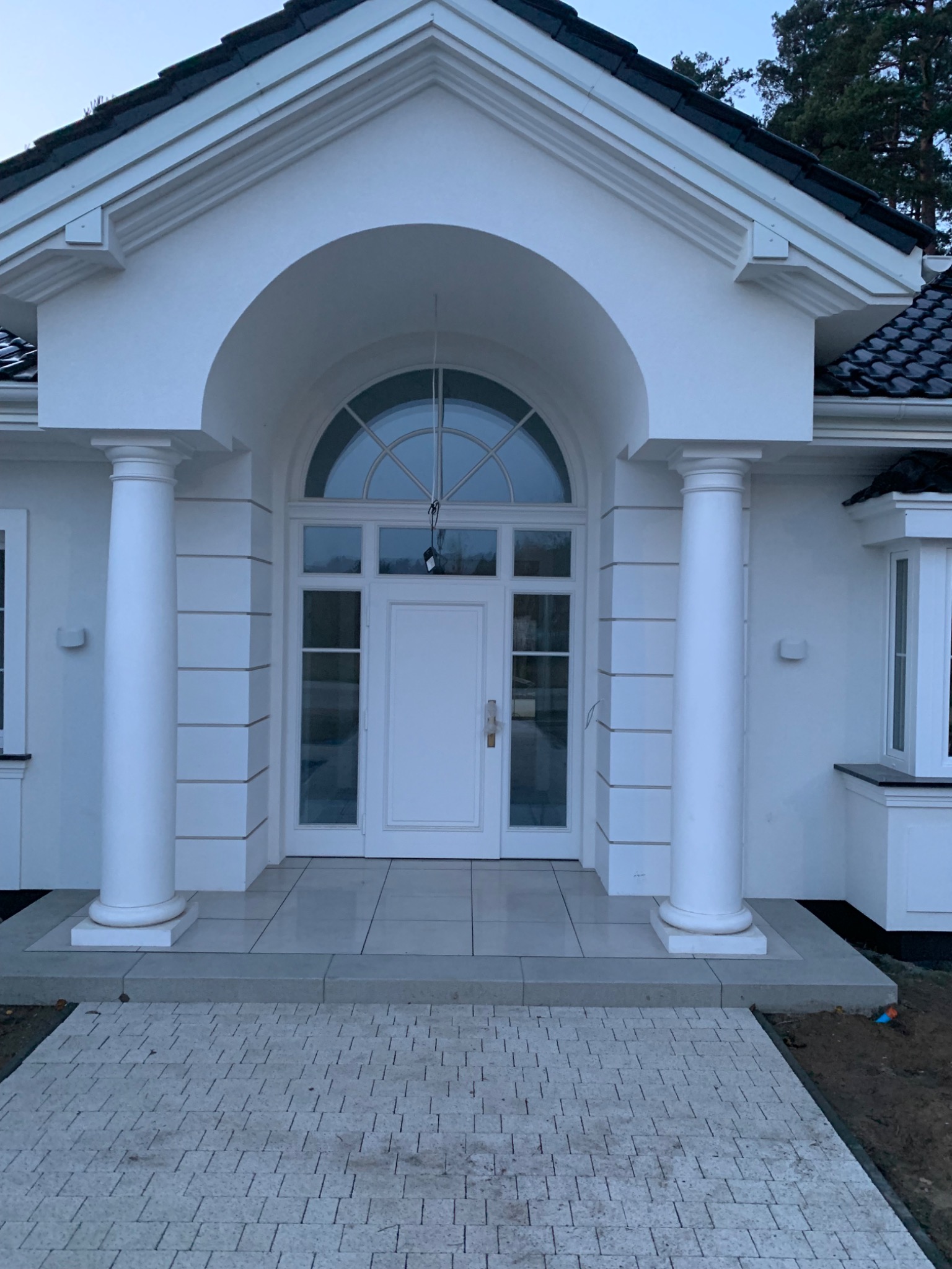 Eleganckie wejście do domu z kolumnami i brukowanym chodnikiem. Biała fasada, łukowe okno nad drzwiami. Nowoczesny bruk w jasnych odcieniach szarości.