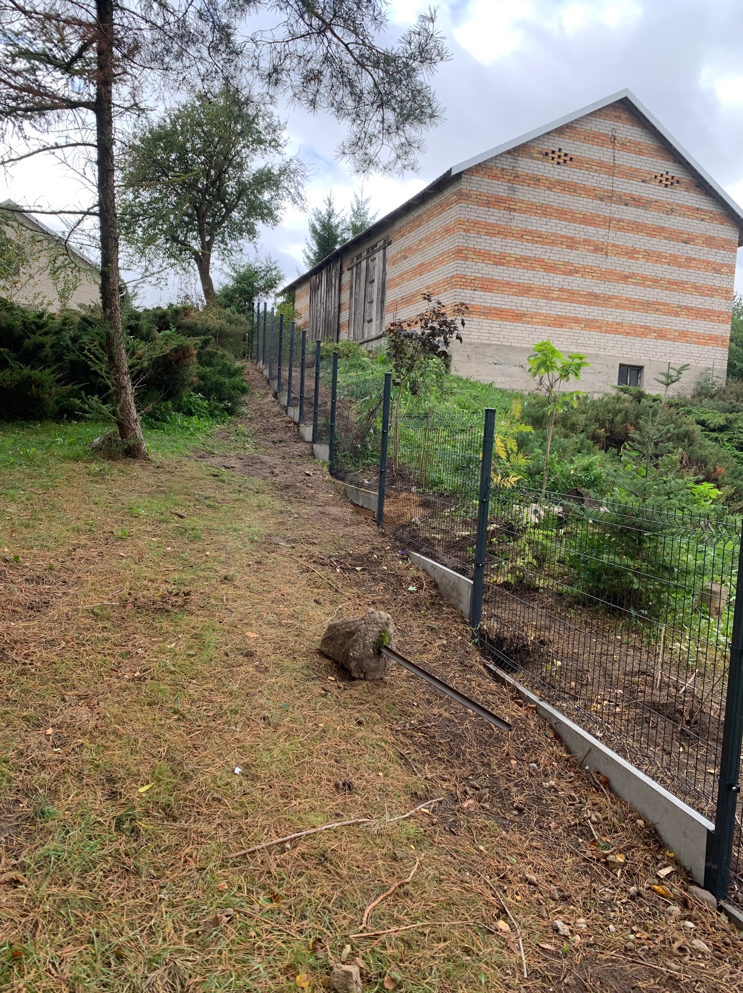 Nowoczesne ogrodzenie panelowe w kolorze grafitowym, biegnące wzdłuż nierównego terenu z widocznym budynkiem w tle. Uwagę zwraca betonowa podmurówka.