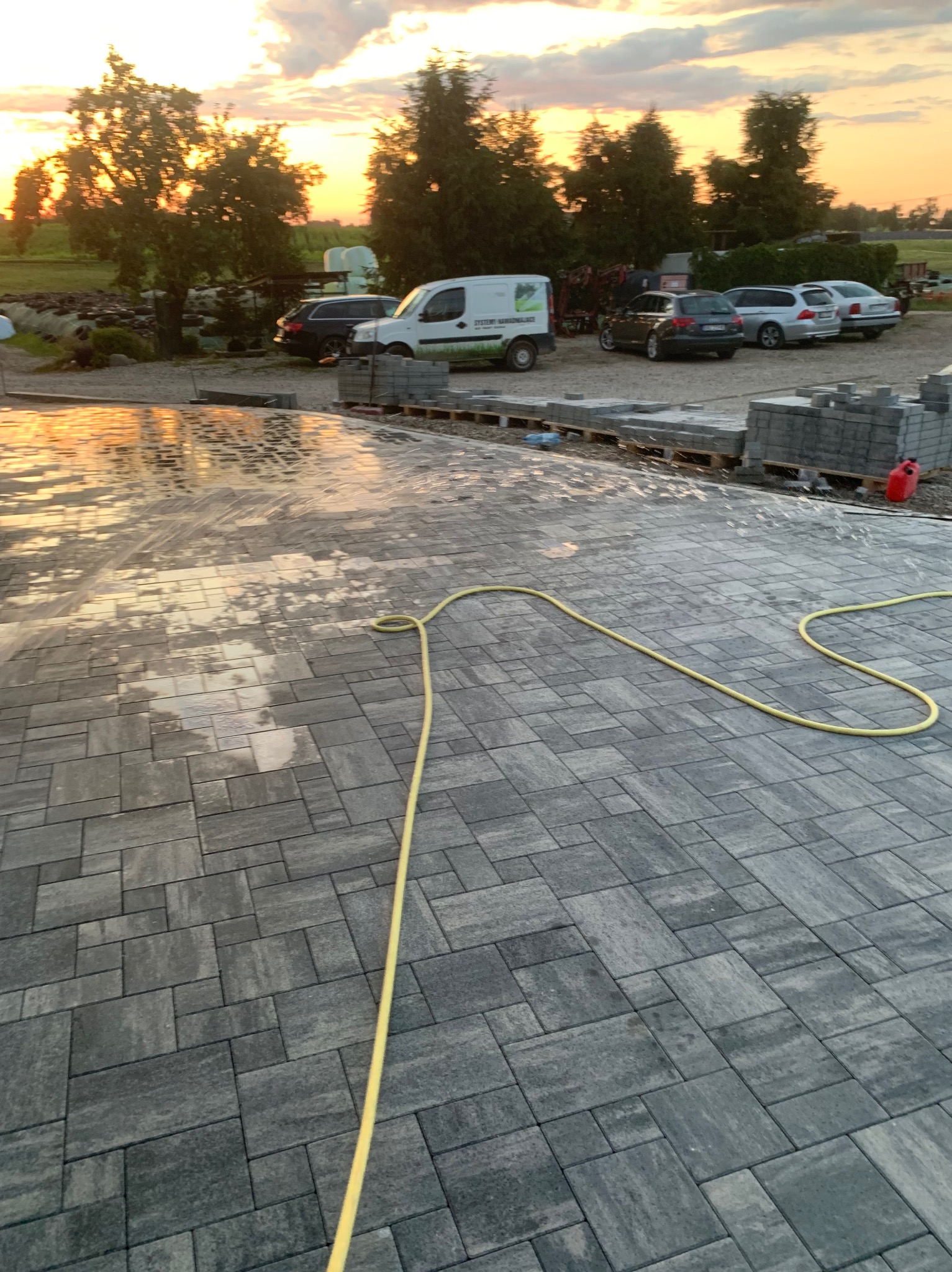 Świeżo ułożona, szara kostka brukowa, częściowo mokra od wody z żółtego węża ogrodowego. W tle zaparkowane auta i zachodzące słońce.