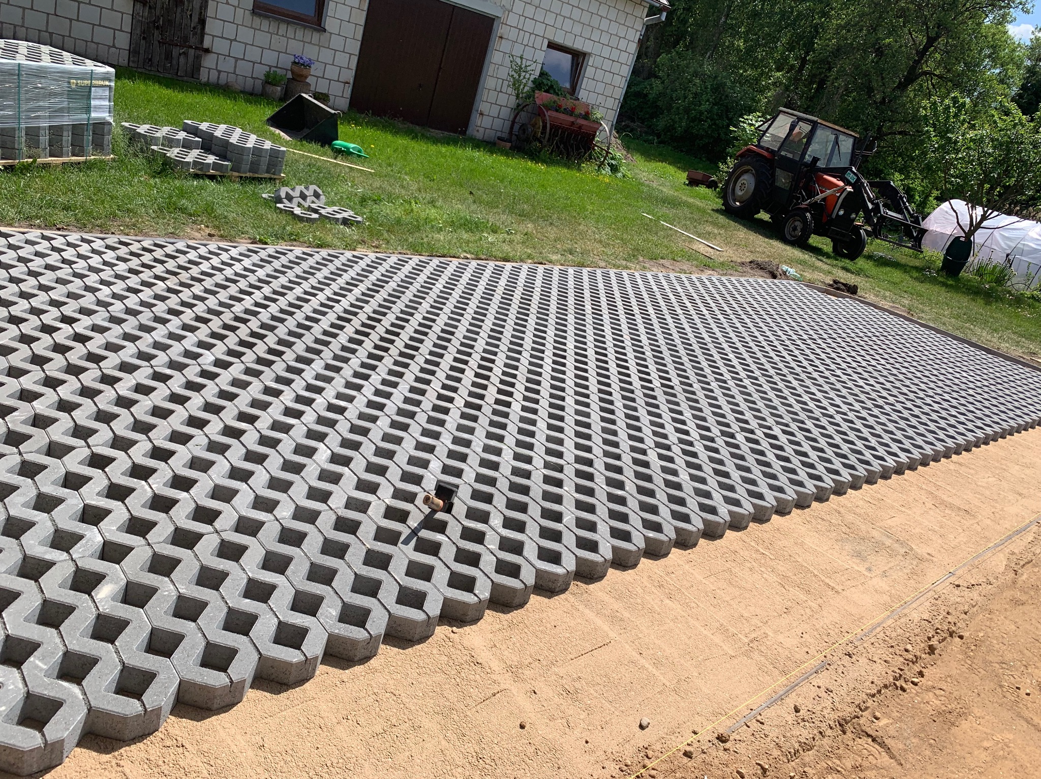 Układanie szarej, ażurowej kostki brukowej na piasku. W tle budynek gospodarczy, traktor i paleta z kostką. Widok z góry, szeroki kadr.