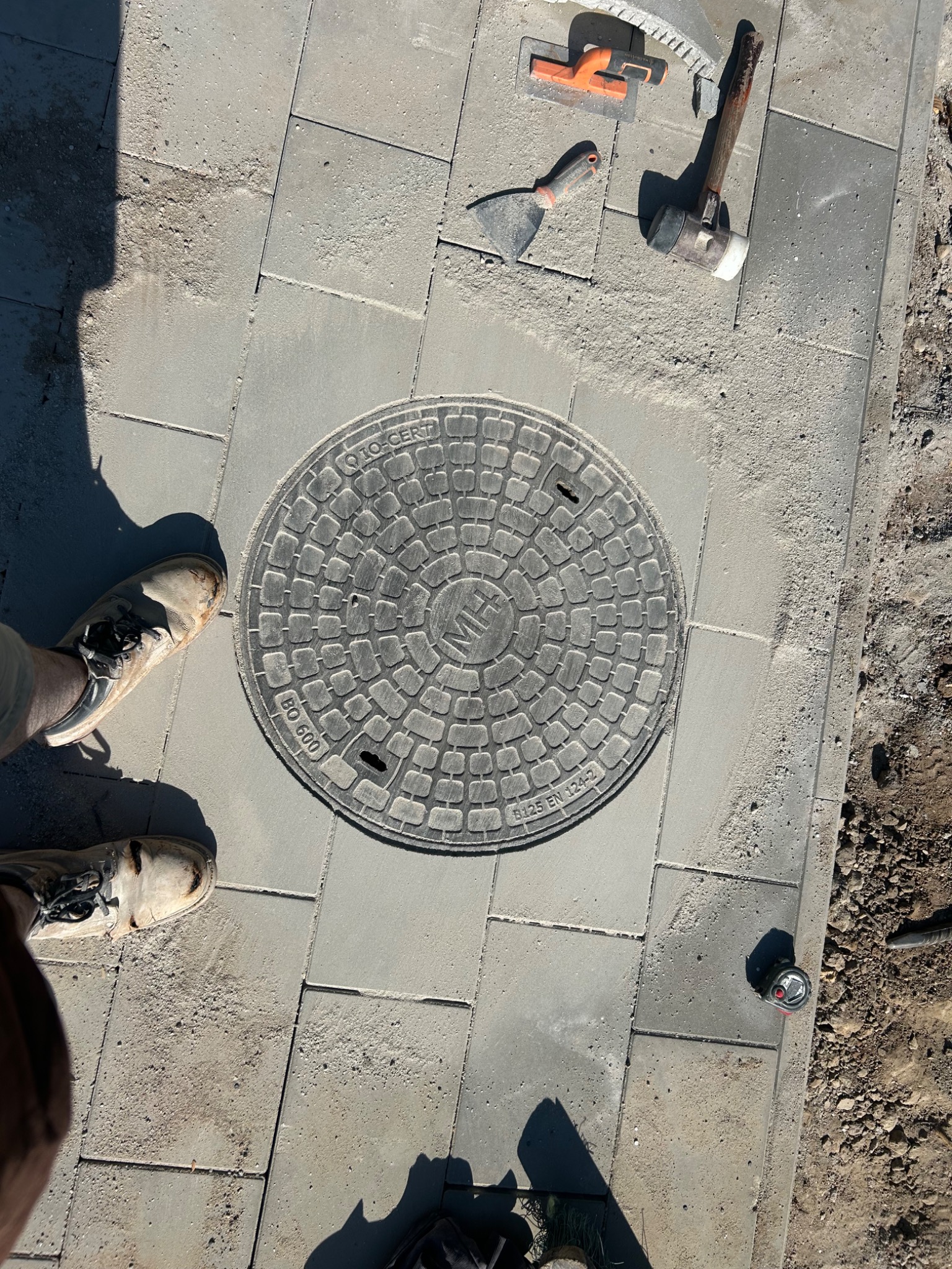 Szare kostki brukowe ułożone wokół studzienki kanalizacyjnej. Widoczne narzędzia brukarskie i fragment buta. Ujęcie z góry, w słoneczny dzień.