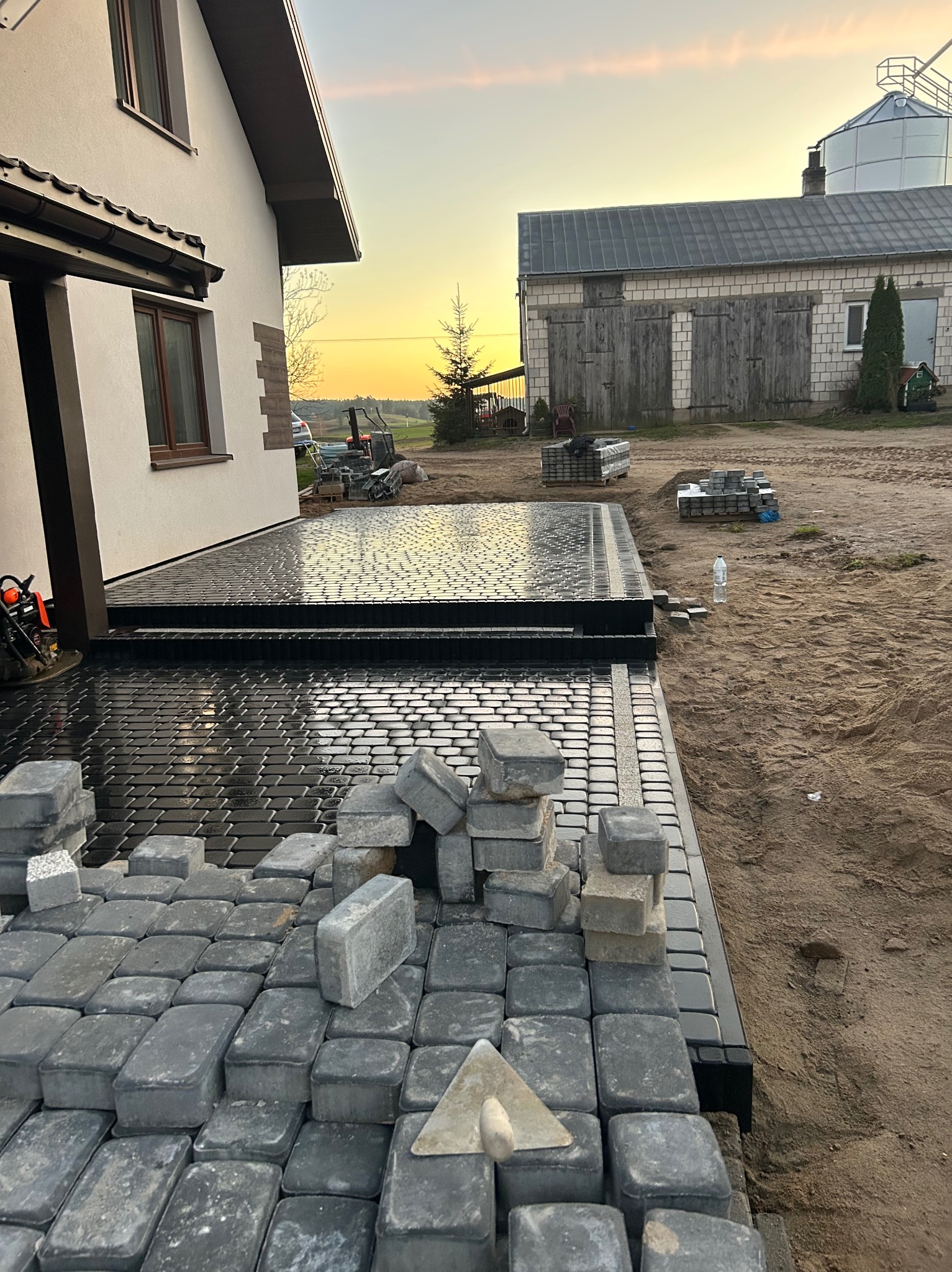 Układanie ciemnoszarej kostki brukowej na posesji domu jednorodzinnego, widoczne narzędzia i materiały budowlane, w tle budynek gospodarczy i zachodzące słońce.