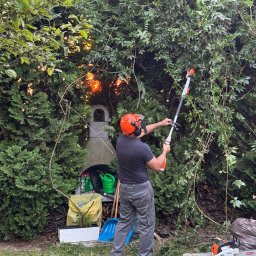 EkoCięcie - Mężczyzna w kasku tnie żywopłot nożycami akumulatorowymi. W tle grill ogrodowy, narzędzia i ścięte gałęzie. Prace ogrodnicze w słoneczny dzień.