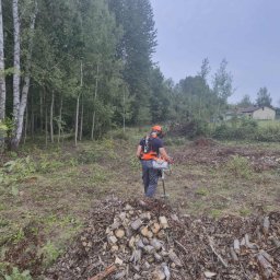 EkoCięcie - Pracownik w kasku i z osprzętem tnącym stoi na terenie po wycince drzew, z widocznym stosem pociętego drewna i pozostałościami po wycince. W tle las i zabudowania.