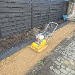 EkoCięcie - Żółta zagęszczarka płytowa na warstwie kruszywa, przygotowująca podłoże pod nawierzchnię, obok ciemnego drewnianego płotu i rabaty ziemi. Widoczna stara kostka brukowa.