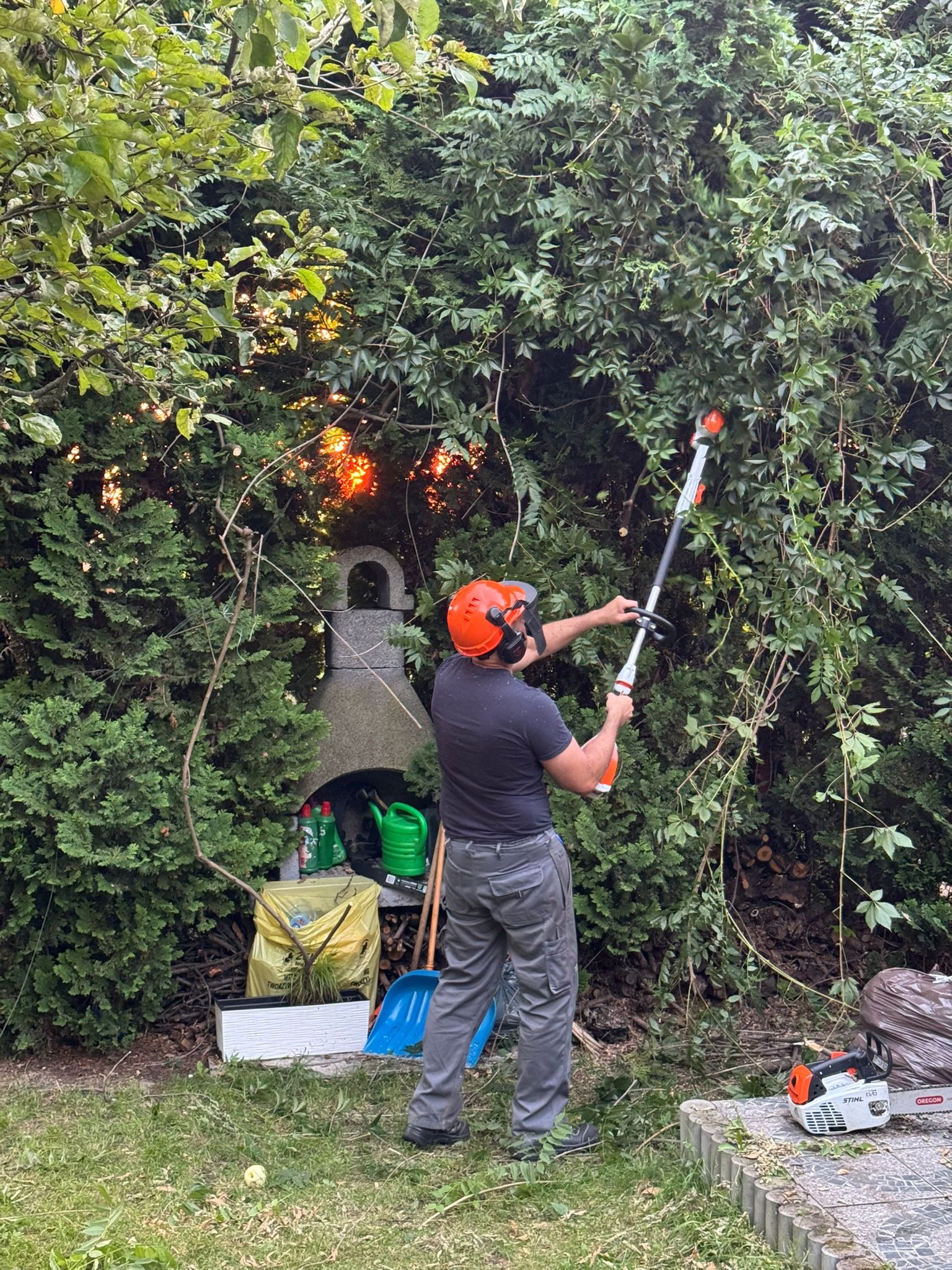 Mężczyzna w kasku tnie żywopłot nożycami akumulatorowymi. W tle grill ogrodowy, narzędzia i ścięte gałęzie. Prace ogrodnicze w słoneczny dzień.