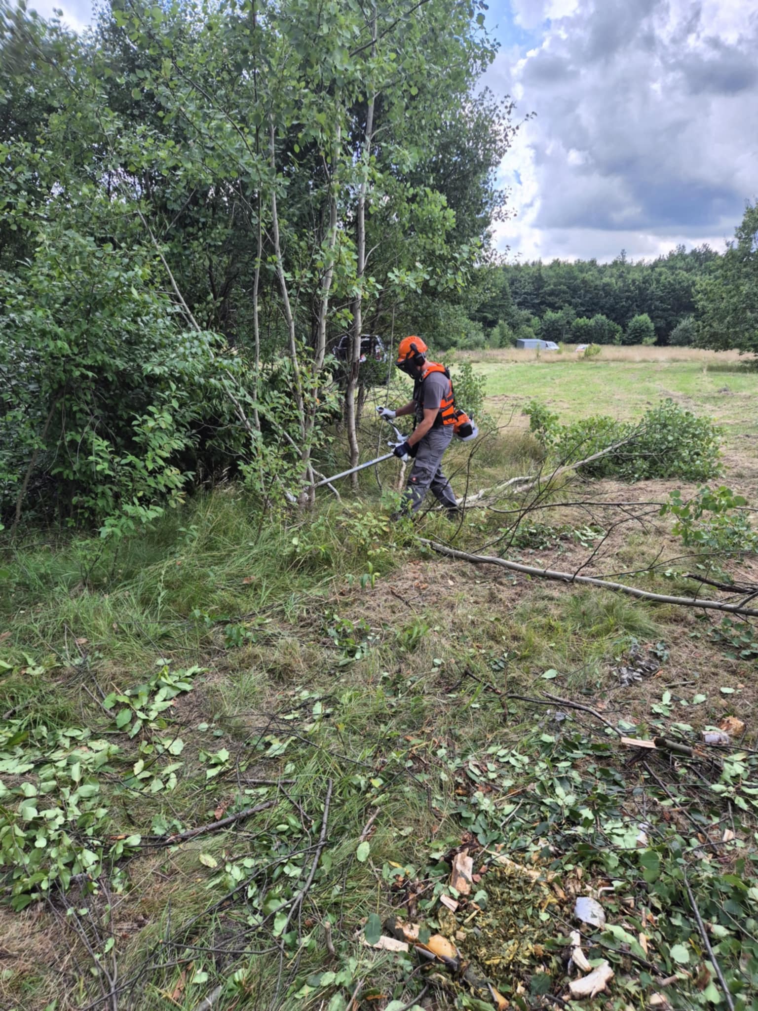 Pracownik w kasku i kamizelce tnie krzaki na zarośniętej łące za pomocą podkaszarki spalinowej. W tle widoczne drzewa i budynek gospodarczy.
