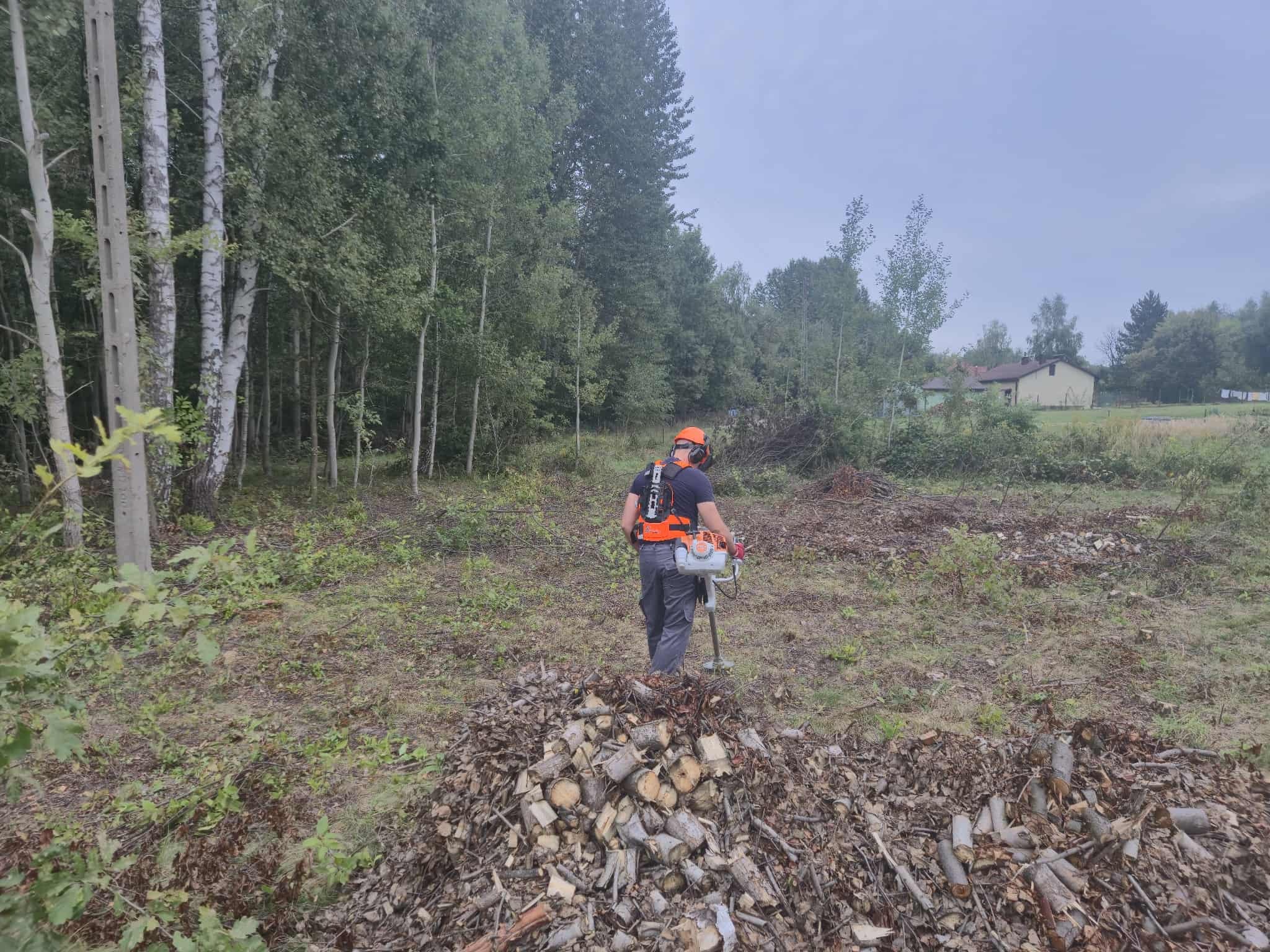 Pracownik w kasku i z osprzętem tnącym stoi na terenie po wycince drzew, z widocznym stosem pociętego drewna i pozostałościami po wycince. W tle las i zabudowania.