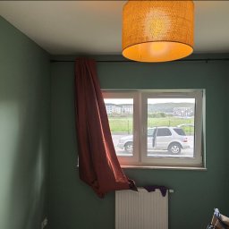 TOMASZ CHOJNACKI - Pokój z zielonymi ścianami, oknem z widokiem na samochód i budynki, oraz pomarańczową lampą. Częściowo zasłonięte okno brązową zasłoną.