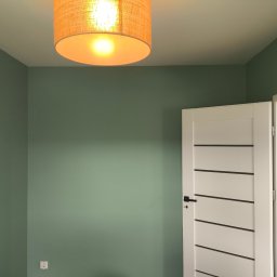 TOMASZ CHOJNACKI - Pokój po malowaniu ścian na oliwkowy odcień, z białymi drzwiami z poziomymi wstawkami i lampą z abażurem w ciepłym odcieniu. Widoczny kontakt elektryczny.