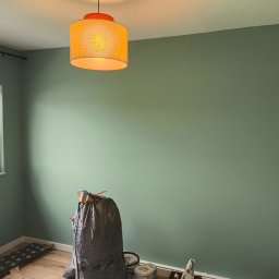 TOMASZ CHOJNACKI - Świeżo pomalowane na zielono ściany pokoju z widocznymi puszkami farby i workiem na śmieci, podkreślone przez ciepłe światło lampy. Widok z dołu.