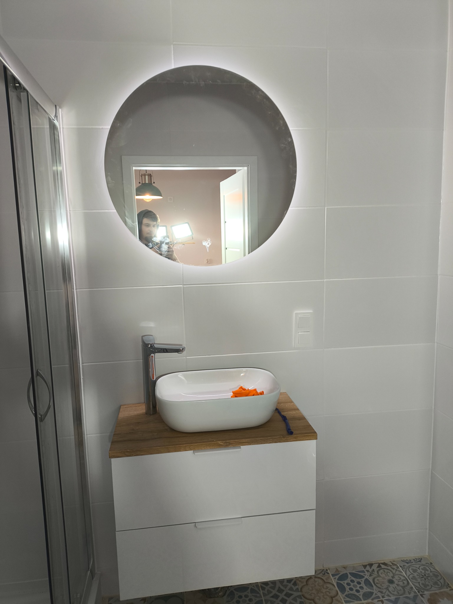 Biała umywalka nablatowa na szafce z drewnianym blatem w łazience z białymi płytkami. W lustrze odbija się osoba z lampą.