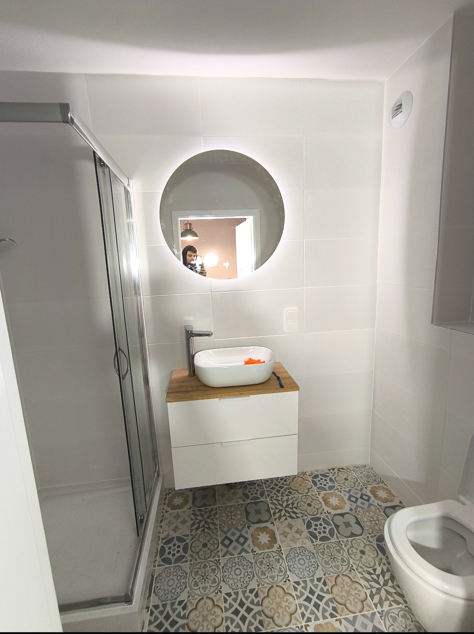 Biała łazienka z kabiną prysznicową i umywalką nablatową, okrągłe lustro z podświetleniem LED. Na podłodze wzorzyste płytki w stylu retro.