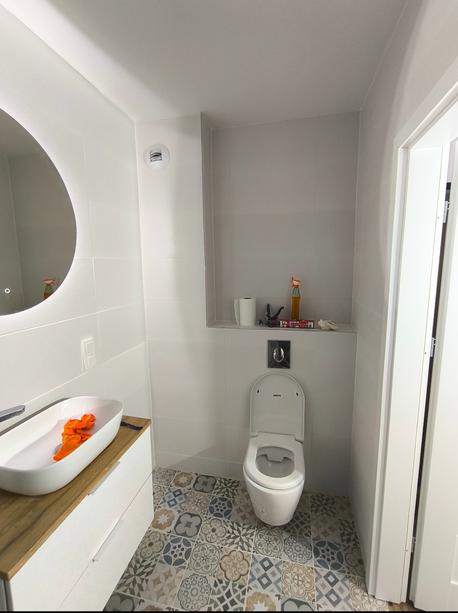 Biała łazienka z okrągłym lustrem, umywalką na szafce z blatem imitującym drewno i toaletą. W tle widoczne narzędzia i akcesoria na półce.