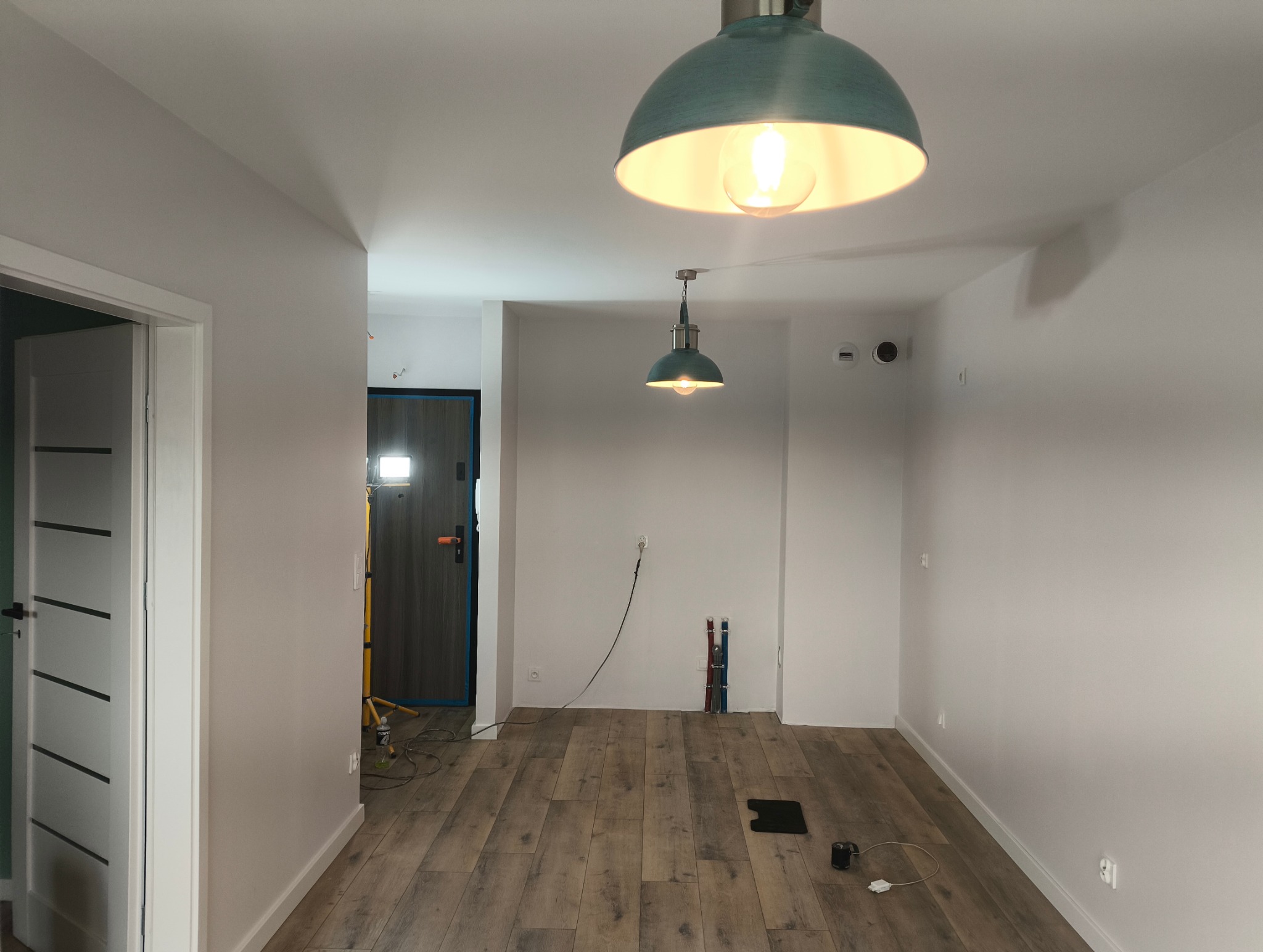 Jasne wnętrze po remoncie z drewnopodobną podłogą, białymi ścianami i turkusowymi lampami. Widoczne przygotowane gniazdka i rury. Drzwi wejściowe w tle.