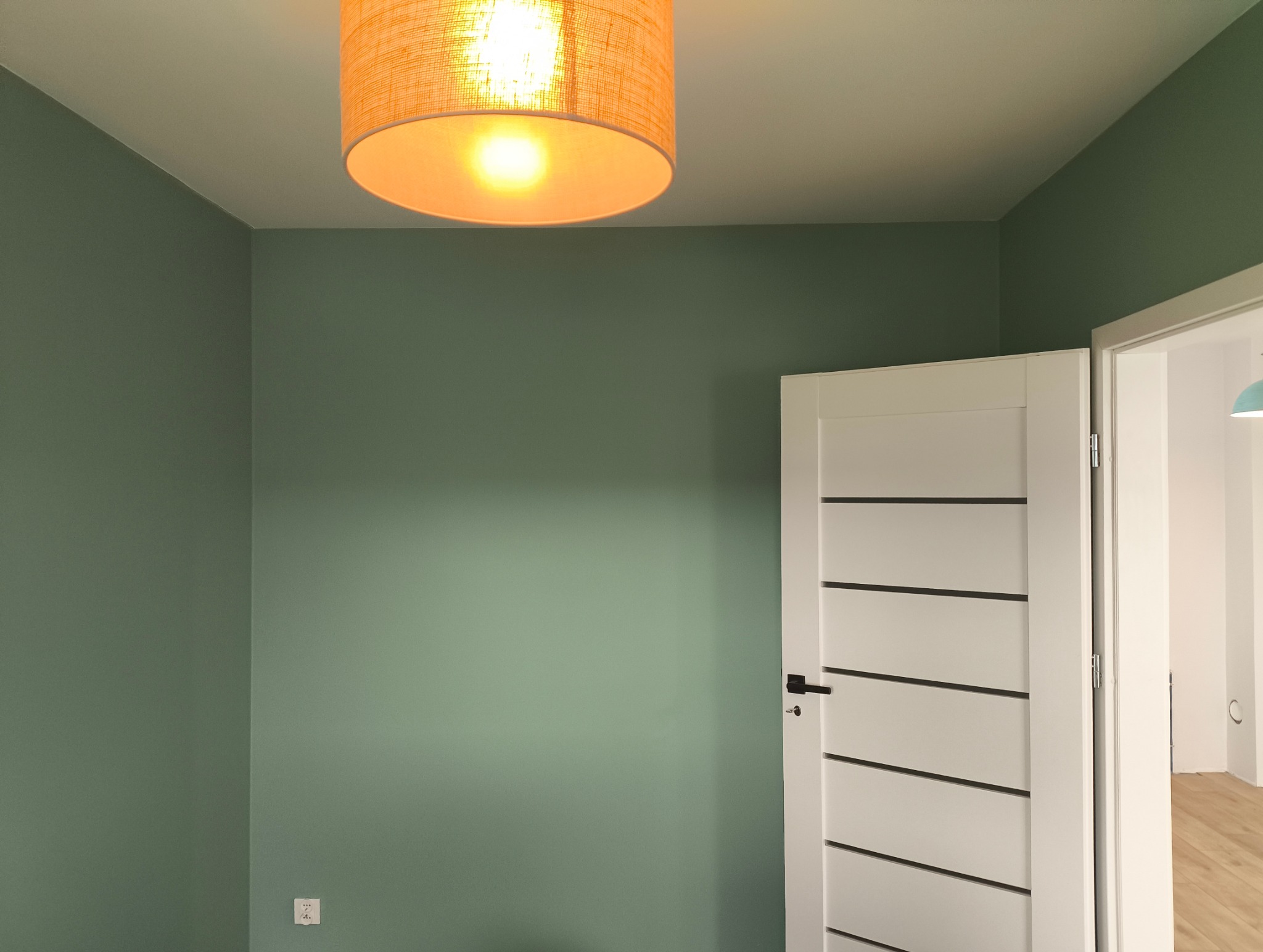 Pokój po malowaniu ścian na oliwkowy odcień, z białymi drzwiami z poziomymi wstawkami i lampą z abażurem w ciepłym odcieniu. Widoczny kontakt elektryczny.