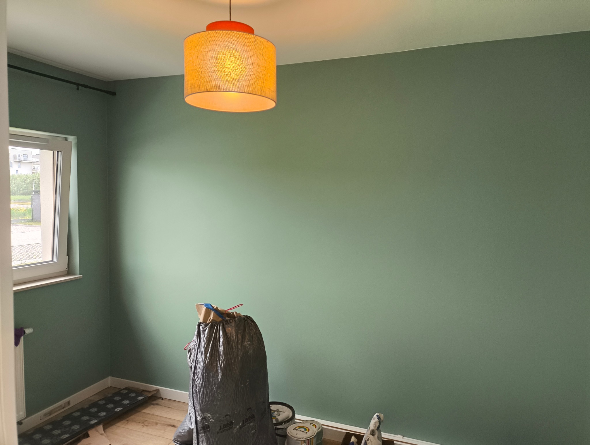 Świeżo pomalowane na zielono ściany pokoju z widocznymi puszkami farby i workiem na śmieci, podkreślone przez ciepłe światło lampy. Widok z dołu.