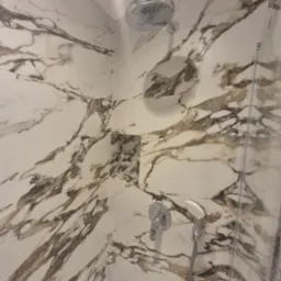 Nowoczesna kabina prysznicowa z marmurowymi płytkami w odcieniach bieli i brązu, chromowana armatura i wnęka na kosmetyki.