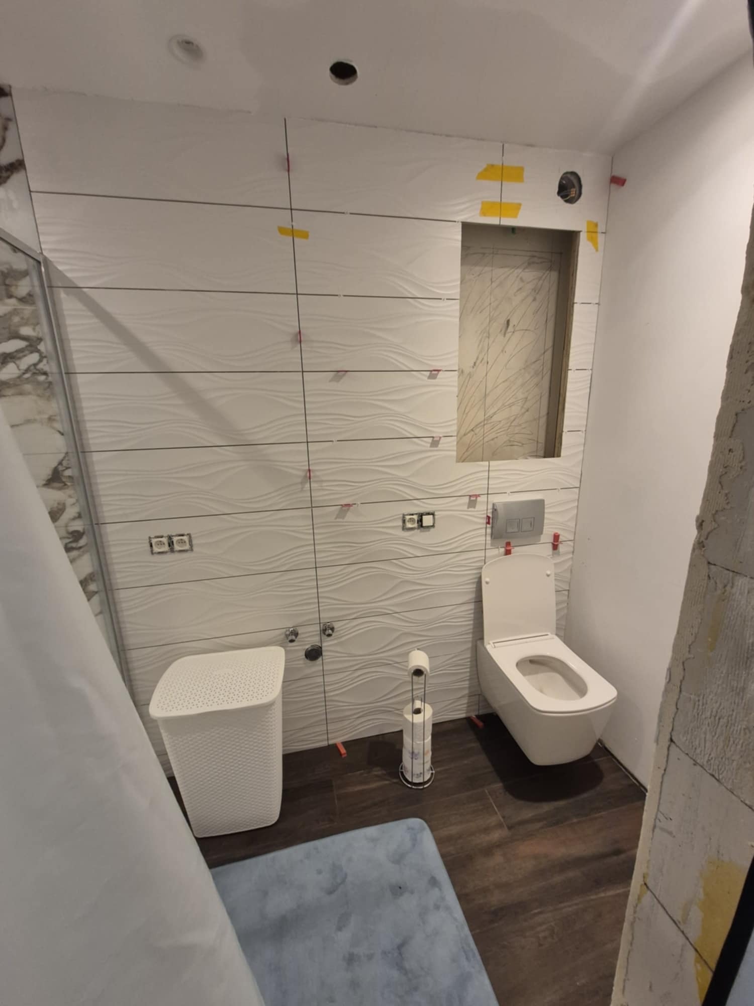 Wnętrze łazienki w trakcie wykańczania: białe, faliste płytki na ścianach, podwieszana toaleta, kosz na pranie. Widoczne elementy montażowe płytek.