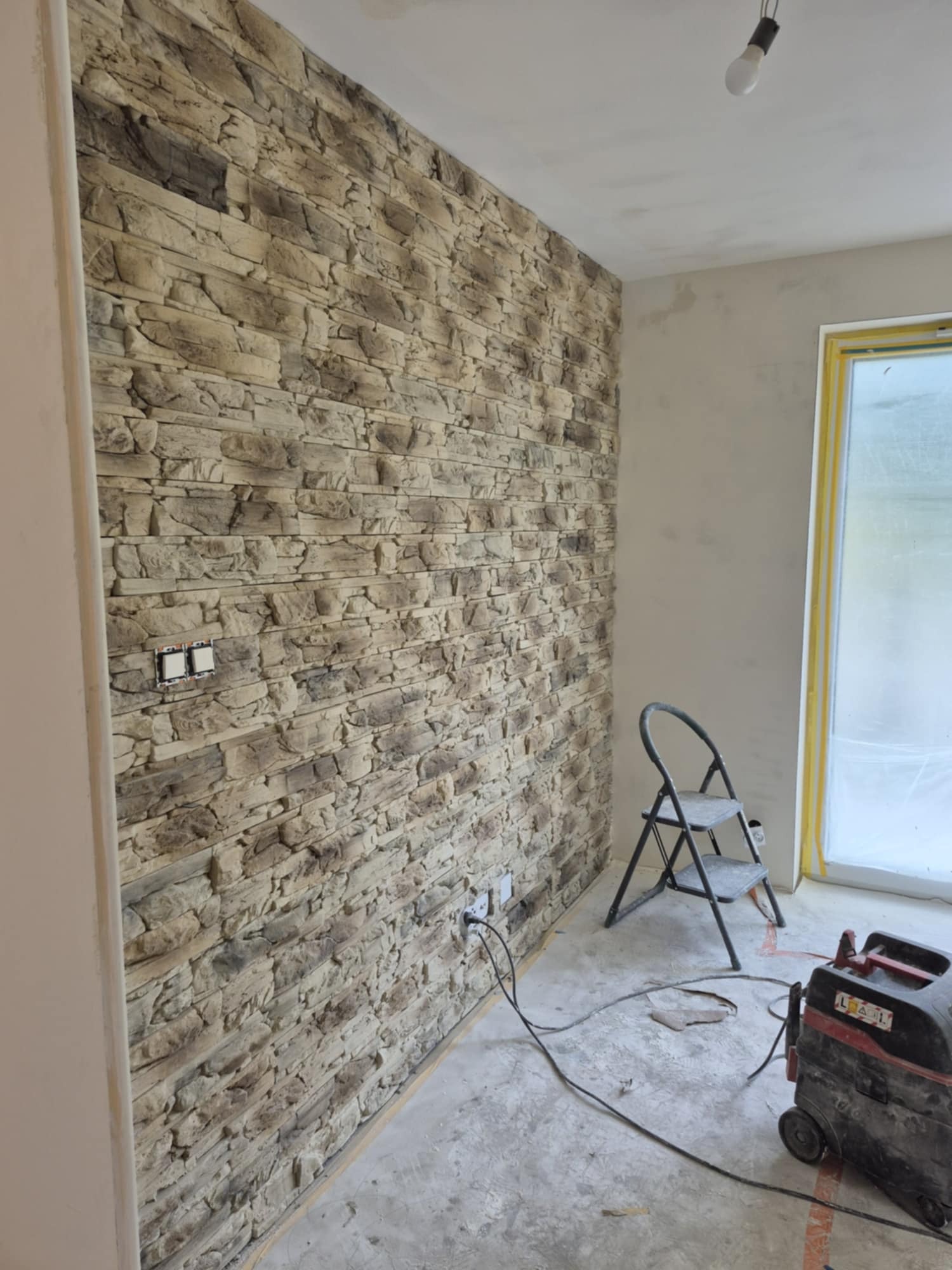 Ściana wykończona kamieniem dekoracyjnym w odcieniach beżu i brązu, widoczne gniazdka elektryczne, drabina i sprzęt budowlany. Wykończenie wnętrz w trakcie realizacji.