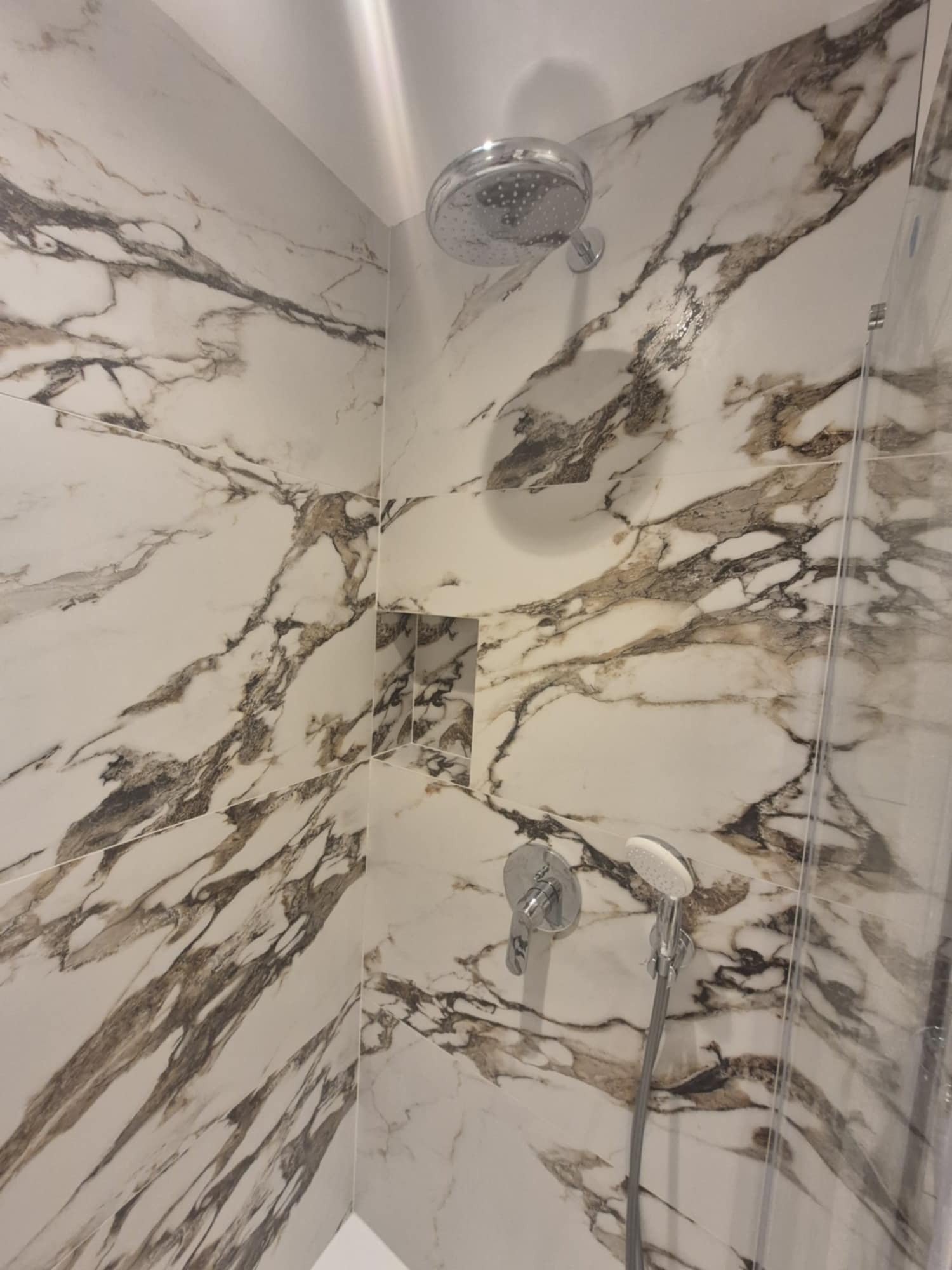 Nowoczesna kabina prysznicowa z marmurowymi płytkami w odcieniach bieli i brązu, chromowana armatura i wnęka na kosmetyki.