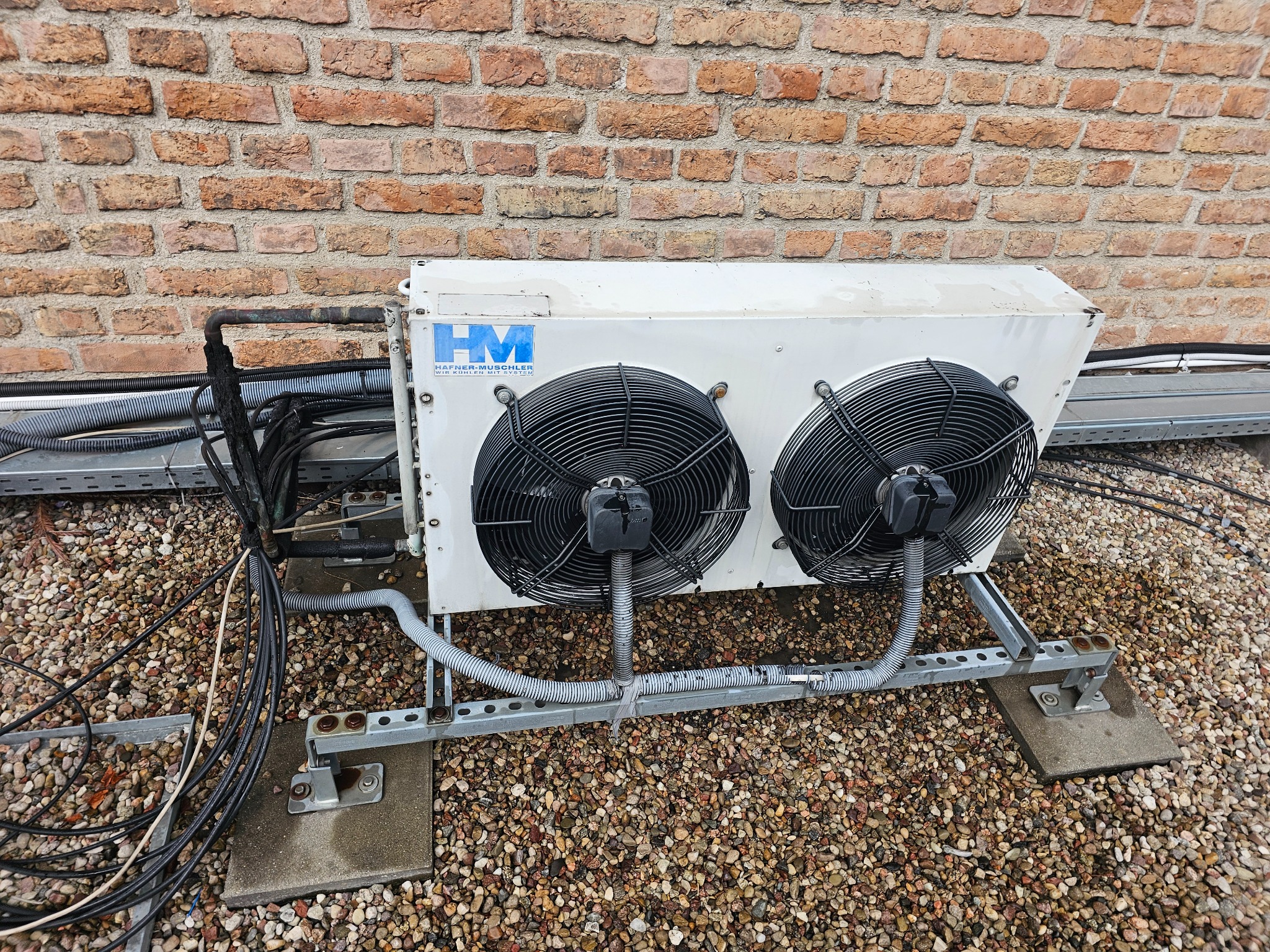 Biała jednostka zewnętrzna klimatyzacji Hafner-Muschler z dwoma wentylatorami na dachu pokrytym żwirem, z widocznymi przewodami i rurami. Tło stanowi ceglana ściana.