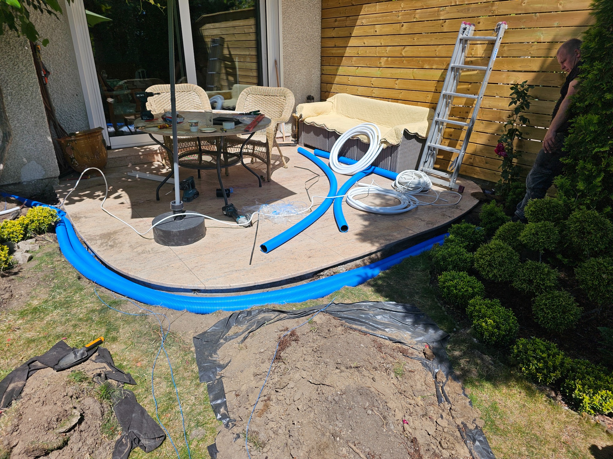 Instalacja niebieskich rur wentylacyjnych na zewnątrz budynku, częściowo zakopanych w ziemi. Na tarasie narzędzia i zwinięte białe kable. Praca w toku.