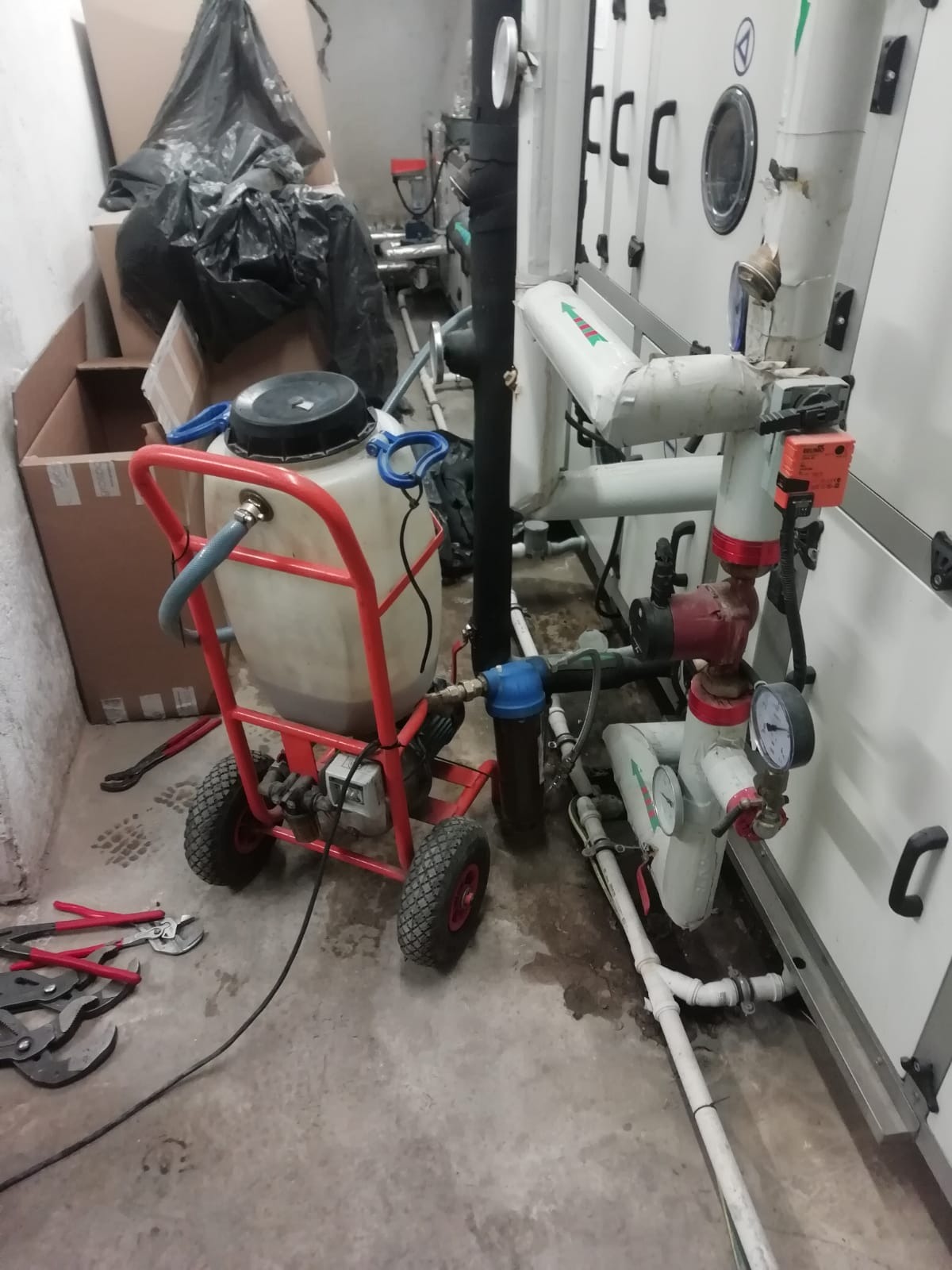 Urządzenie do czyszczenia instalacji grzewczej na wózku z zbiornikiem, obok rur i armatury w pomieszczeniu technicznym. Widoczne narzędzia.