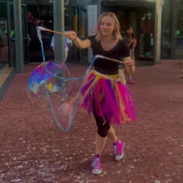 Animator w kolorowej spódniczce tworzy gigantyczne bańki mydlane w plenerze, na tle przeszklonego budynku. Dziewczynka obserwuje pokaz.