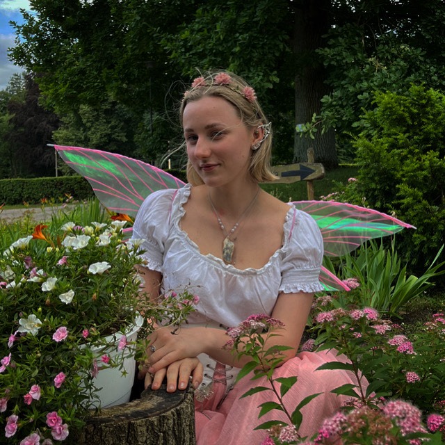 Portret młodej kobiety w kostiumie wróżki z różowymi skrzydłami, siedzącej wśród kwiatów na tle zieleni. Na głowie ma wianek, a na uszach spiczaste nakładki.