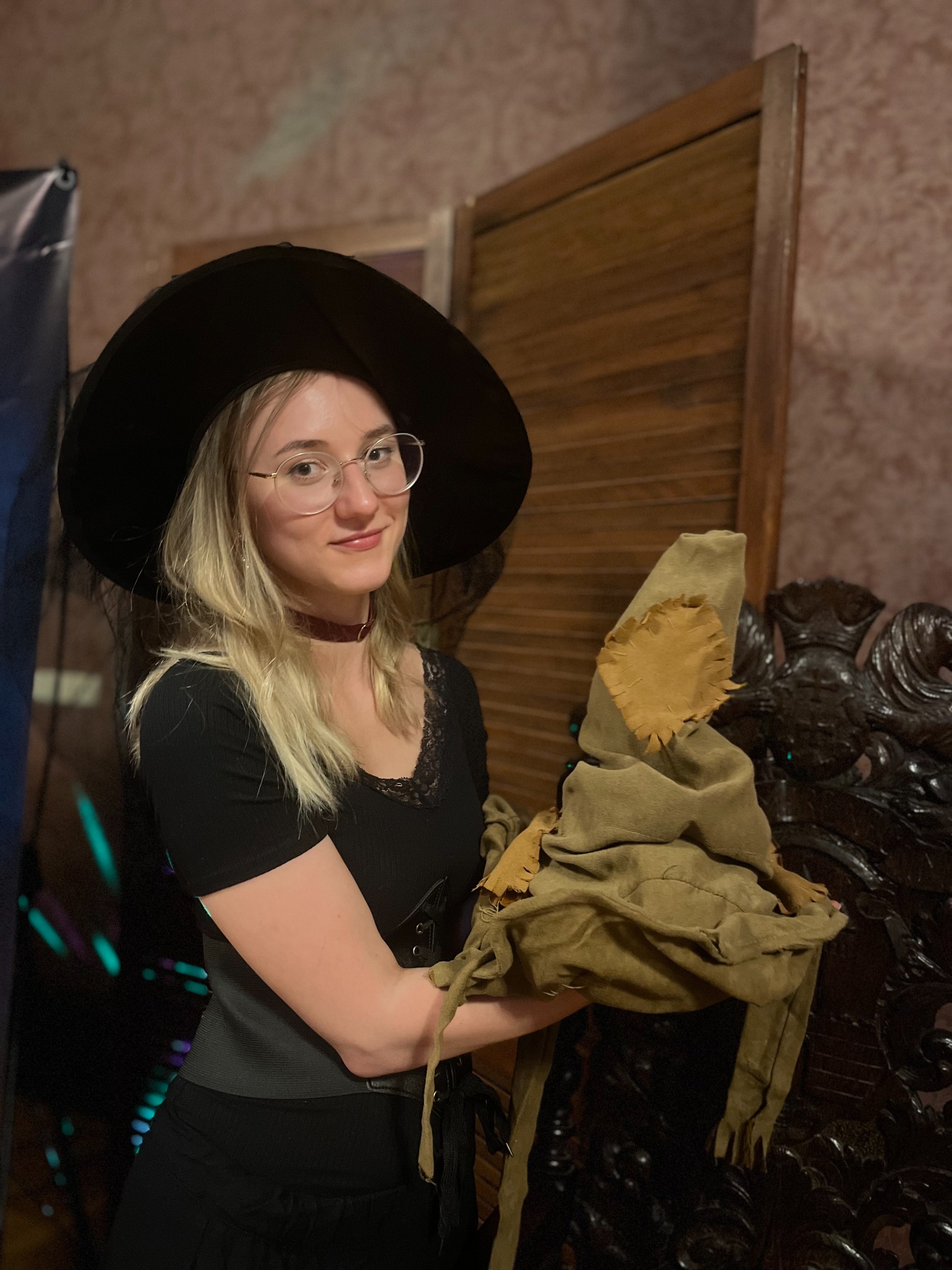 Uśmiechnięta blondynka w okularach i kapeluszu czarownicy trzyma brązowy kapelusz z łatą na tle drewnianej ściany i dekoracji imprezowych.