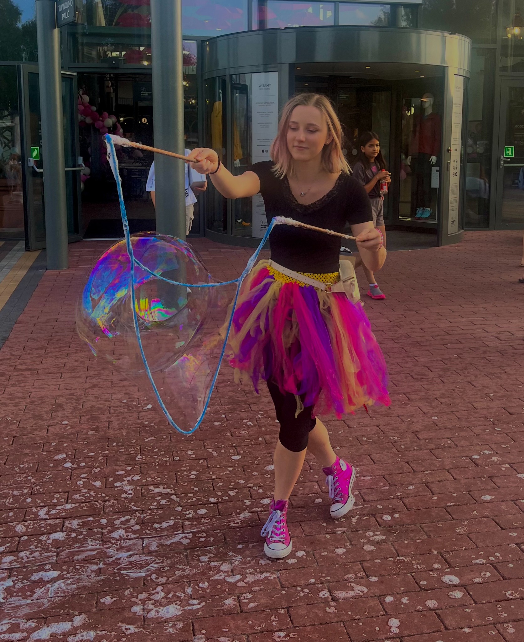 Animator w kolorowej spódniczce tworzy gigantyczne bańki mydlane w plenerze, na tle przeszklonego budynku. Dziewczynka obserwuje pokaz.