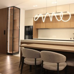 Nowoczesna kuchnia od 10na10 Studio łączy minimalizm i funkcjonalność. Zabudowa w drewnie i kaszmirze z blatami z konglomeratu oraz wyspą i stołem tworzy spójną aranżację, a designerska lampa podkreśla elegancję wnętrza.