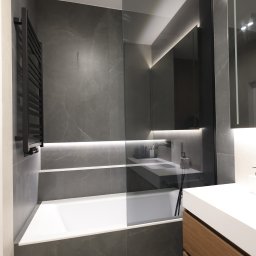 Łazienka od 10na10 Studio utrzymana w eleganckiej szarości z drewnem i bielą. Zabudowa z forniru, wanna z przeszkleniem i podświetlane lustro tworzą nowoczesną aranżację. Czarne detale armatury podkreślają minimalistyczny charakter wnętrza.