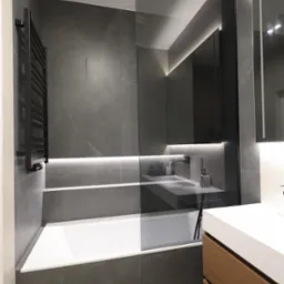 Elegancka szara łazienka z drewnianymi akcentami. Wanna z przeszkleniem, podświetlane lustro i czarna armatura tworzą minimalistyczny, nowoczesny wygląd.