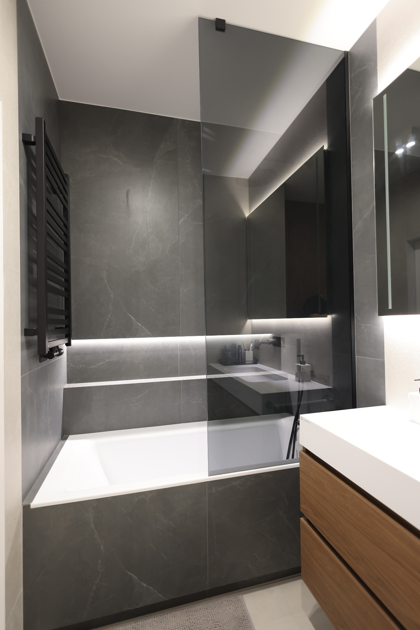 Elegancka szara łazienka z drewnianymi akcentami. Wanna z przeszkleniem, podświetlane lustro i czarna armatura tworzą minimalistyczny, nowoczesny wygląd.